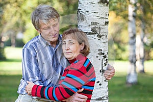 Elderly man embraces his wife and birch tree, looking at woman, half length portrait