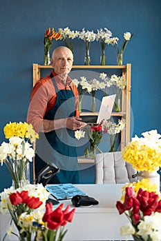 An elderly man of European appearance, a salesman-florist, smiles confidently while using a laptop in a flower shop