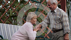 Elderly Man Gives Hand To His Wife Helping Her Get Up From The Bench At Park
