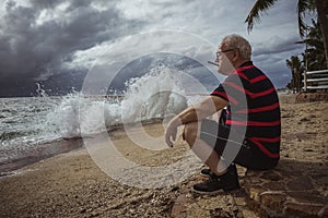 Elderly man in glasses and a striped t-shirt sitting on the beach and looking at the storm smokes a cigar. A wise man in