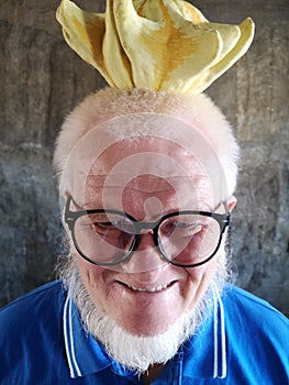 An elderly man with glassesÑŒ and a gray beard in a blue t shirt with a yellow decorative pumpkin on his head poses for the camera
