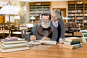 Elderly man grandfather and his granddaughter reading exciting book together in vintage old library, on the background