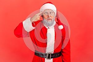 Elderly man with gray beard in santa claus costume crossed eyes, showing stupid gesture, looking at camera with condemnation and