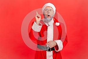 Elderly man with gray beard wearing santa claus costume raising finger and having genius idea of