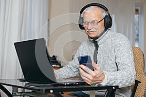Elderly man with headphones works behind modern laptop. old man holds phone with blank screen. Senior man shopping in online store