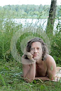 An elderly man with lies sunbathes in the grass on the shore. Russian, long hair, brunette.