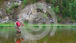 An elderly man with long hair and a santa claus beard stands in the water and holds a fishing rod. Fly fishing