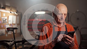Elderly man mechanic in a bicycle repair shop using his mobile phone reading a text message, working in workshop or