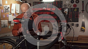 Elderly man mechanic chatting on phone, reading social media, asking for help on repairing damaged bike in his workshop