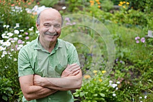 An elderly man with a mustache and a bald spot in a green T-shirt is standing among flowers in the summer garden, arms