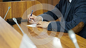 An elderly man - politician, lawyer or teacher writes on a piece of paper while sitting at a table in the boardroom. A court