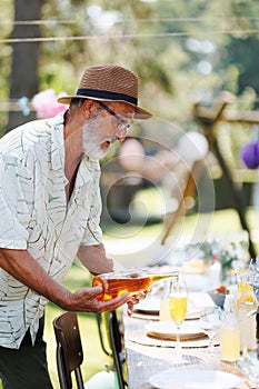 Elderly man preparing refreshments for a summer garden party, pouring wine in glass.