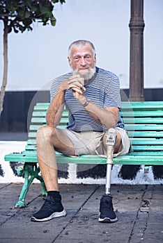 Elderly man with prosthetic leg. left rests sitting on a park bench. Spending a relaxing afternoon. People concept