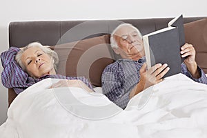 Elderly man reading book while spouse sleeping besides in bedroom