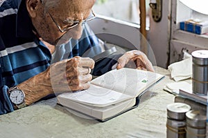 Elderly man reading writings in a notebook with magnifying glass near the window at home