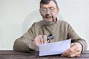 Elderly man reads fine print on white piece of paper using magnifying glass. Magnifier helps old senior