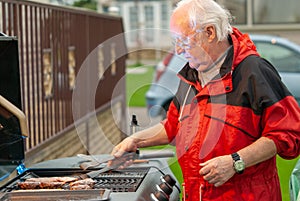 Elderly man in a red coat cooking meat on a BBQ