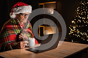 Elderly man in Santa hat holding cup of cacao while watching movie film on laptop at home for Christmas. New Year and
