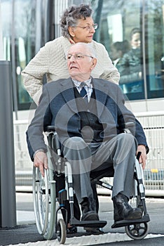 Elderly man seated in wheelchair pushed by wife