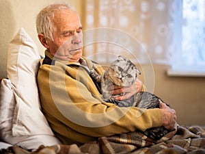 Elderly man sits on a bed with a scottish fold cat in his arms