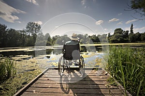 Elderly man sits at the edge of a calm lake,wearing a panama hat,sitting relaxed in his wheelchair.Andover,Hampshire,United