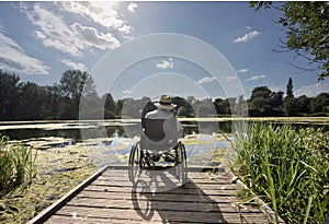Elderly man sits at the edge of a calm lake,wearing a panama hat,sitting relaxed in his wheelchair.Andover,Hampshire,United