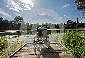 Elderly man sits at the edge of a calm lake,wearing a panama hat,sitting relaxed in his wheelchair.Andover,Hampshire,United