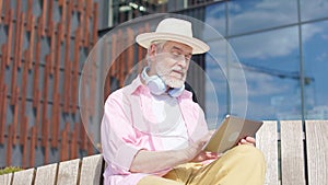Elderly man sitting on bench and focusing on tablet in hands