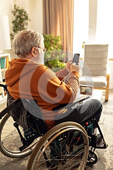 Elderly man sitting on wheelchair in living room at home and using mobile phone for communication, leisure and news