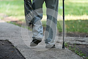 An elderly man slowly walks along the path leaning on a cane. In his hand he holds a plastic bag with the purchased products