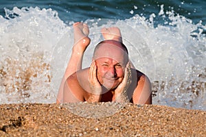 elderly man squints from the sun lying on the sand in the sea foam and basking in the waves