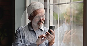 Elderly man standing indoor lean on window using smartphone device