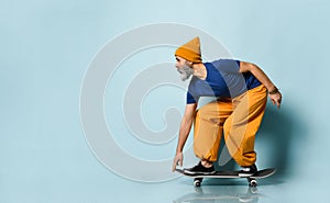 Elderly man in t-shirt, orange pants, hat, gumshoes. Riding black skateboard, posing sideways on blue background. Full length