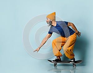 Elderly man in t-shirt, orange pants, hat, gumshoes. Riding black skateboard, posing sideways on blue background. Full length