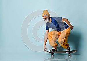 Elderly man in t-shirt, orange pants, hat, gumshoes. Riding black skateboard, posing sideways on blue background. Full length