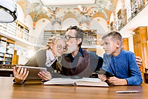 Elderly man teacher and his teen students, boy and girl, sitting at the table in library, reading book, using tablet and