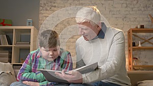 Elderly man teaches a young fat guy how to read. Grandfather and grandson, happy family, home cosiness concept. 60 fps