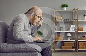 Elderly man touching his glasses rim looking attentively at laptop computer