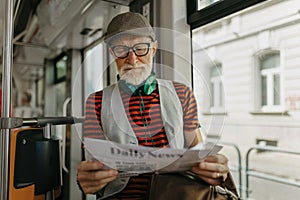 Elderly man traveling through the city by bus, reading newspaper. Senior city commuter taking tram to grocery, using