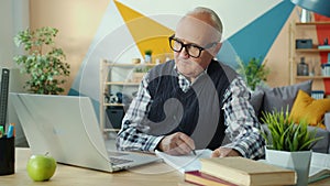 Elderly man using laptop computer typing going online taking notes at home