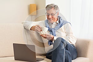 Elderly Man Using Laptop Having Coffee Sitting On Couch Indoor