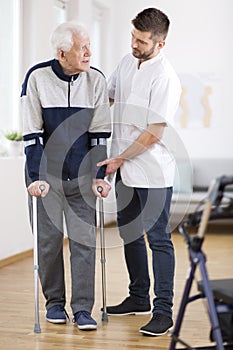 Elderly man walking on crutches and a helpful male nurse supporting him