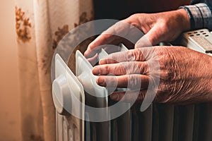 An elderly man warms his hands over an electric heater. In the off-season, central heating is delayed