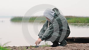 An elderly man warms his hands over a smoldering fire on the shore of the reservoir in autumn.