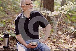 Elderly man wearing glasses enjoys a cup of tea while sitting on a fallen tree trunk