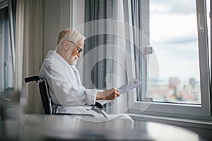 Elderly man in a wheelchair reading the newspaper in his robe in the morning.