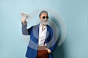 Elderly man in white shirt, jacket, brown pants and sunglasses. He showing fan of hundred dollar bills, posing on blue background