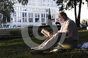 An elderly man in a white shirt is sitting on a blanket, on the ground in a park and reading an interesting book. A