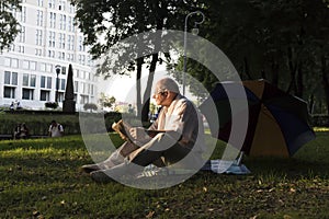 An elderly man in a white shirt is sitting on a blanket, on the ground in a park and reading an interesting book. A