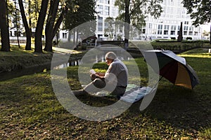 An elderly man in a white shirt is sitting on a blanket, on the ground in a park and reading an interesting book. A